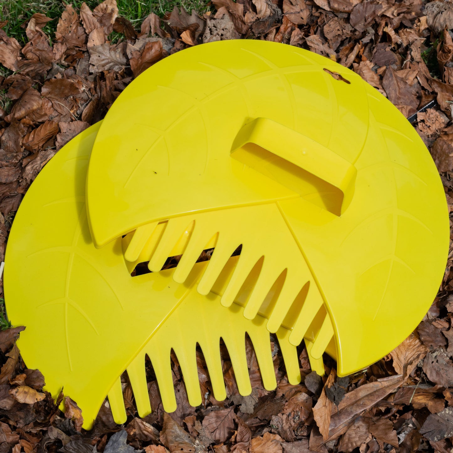 Yellow leaf grabber garden scoop resting on leaves, with front grip and rake teeth