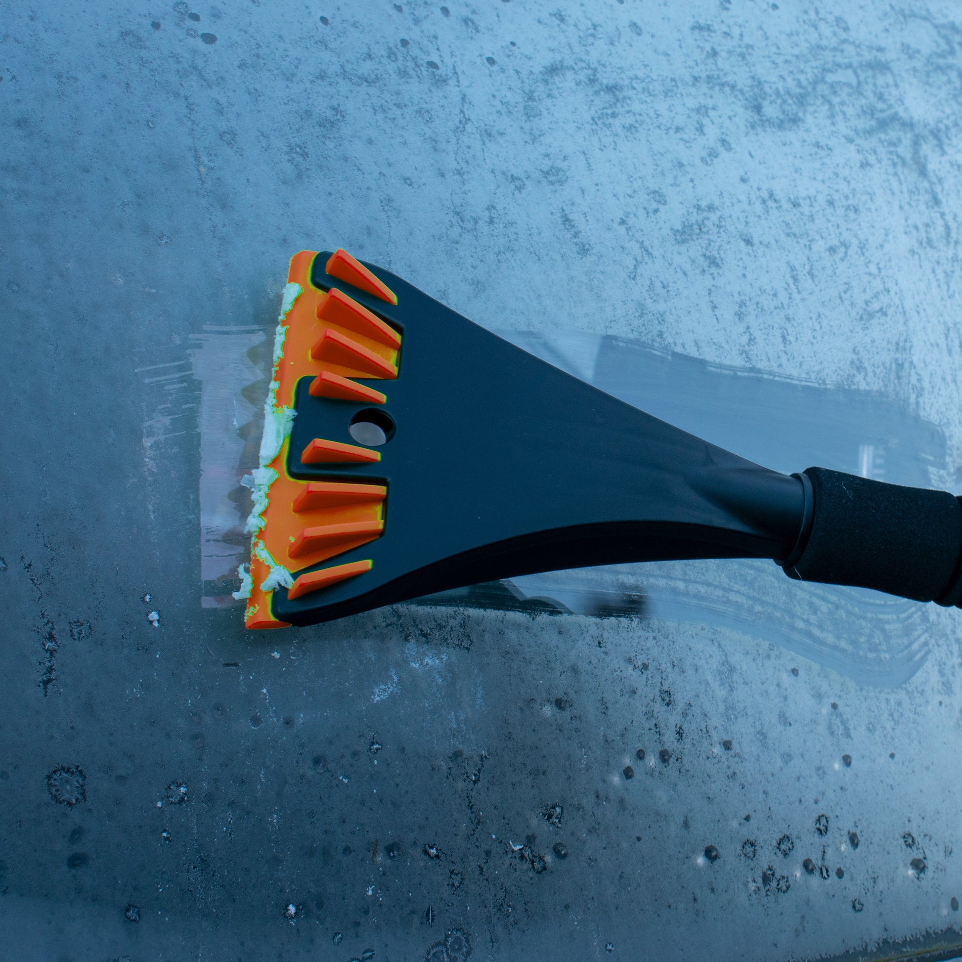 Orange bristle snow brush and scraper blade clearing frost from car window